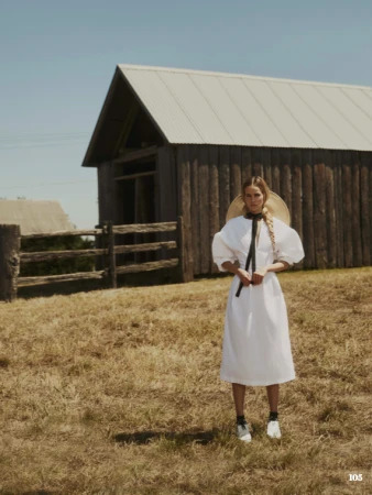 Woman in a white dress and straw hat stands in a field near a wooden barn under a clear blue sky on a sunny day.