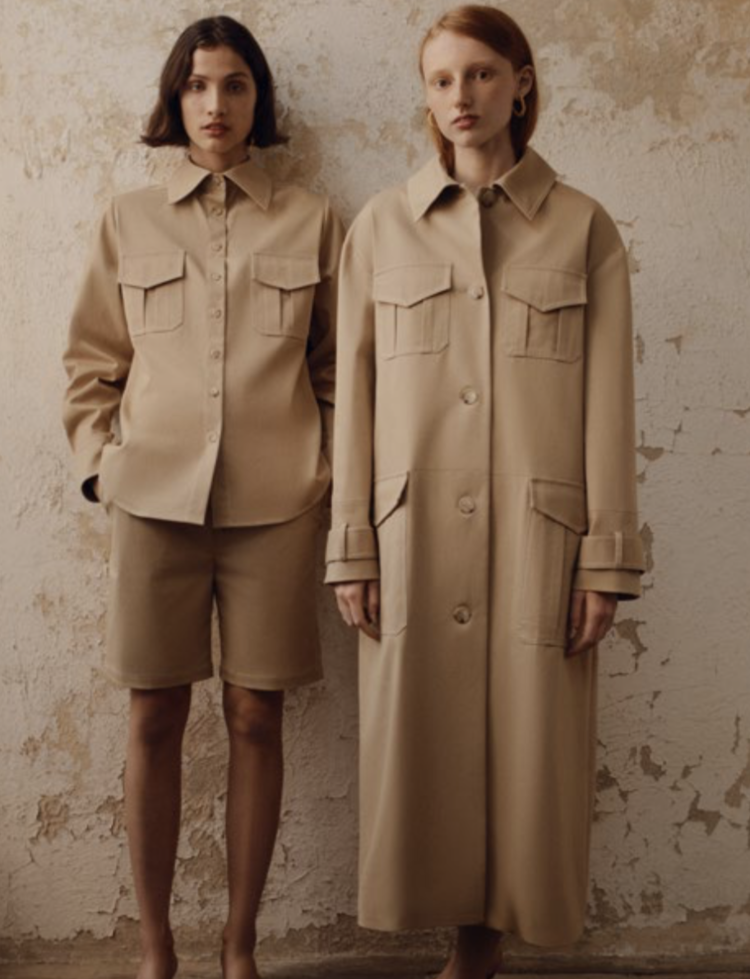Two adult women pose indoors against a mottled beige wall. One wears a short set, the other a long trench coat, both in matching beige.