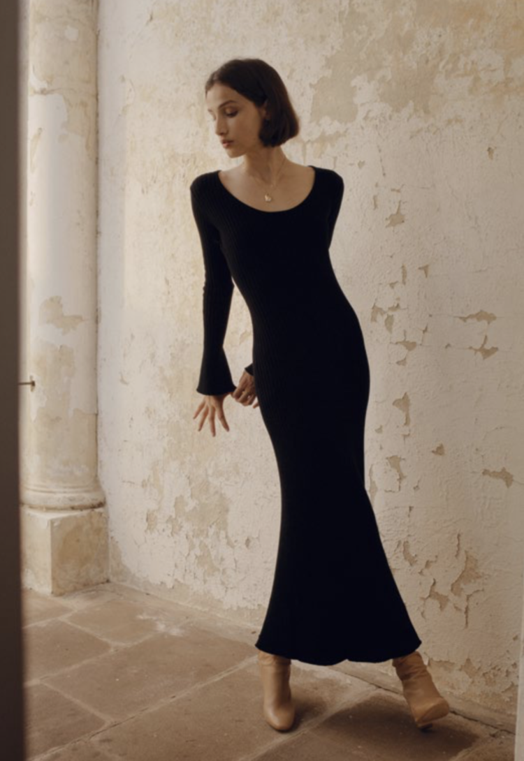 An adult female poses indoors wearing a long-sleeved black dress and tan boots, standing in front of a distressed textured wall.