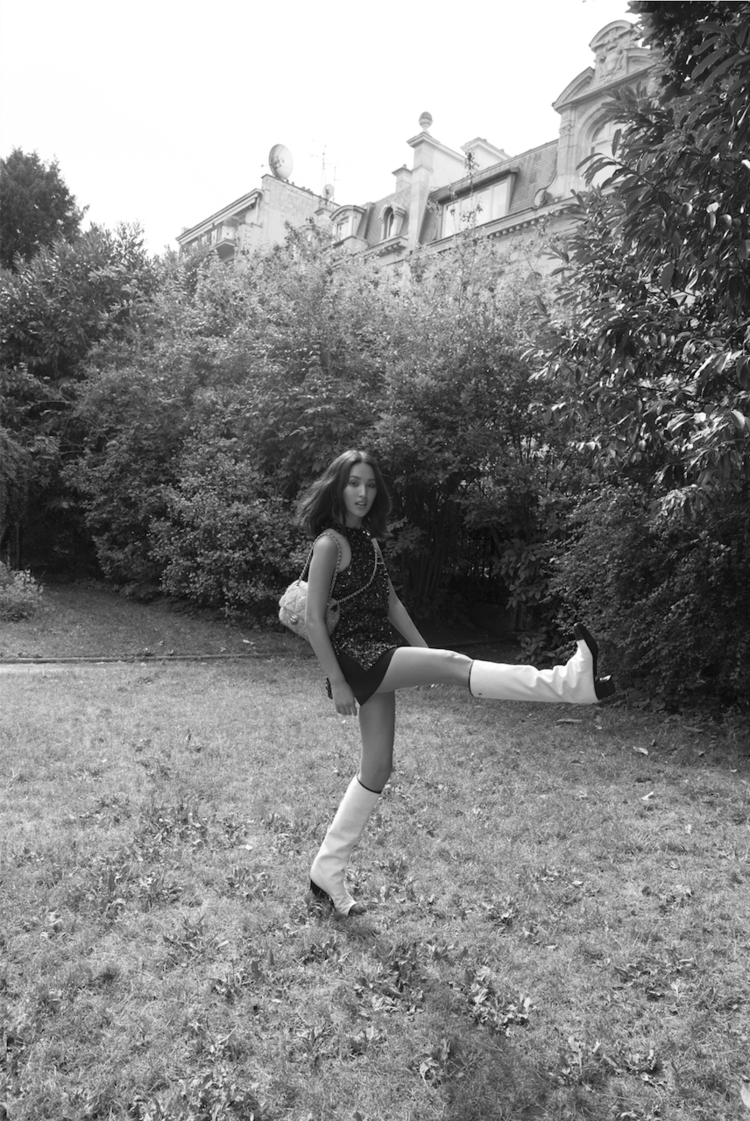 Woman in mini dress and white boots poses in a kicking stance outside near a building and trees on a grassy field in black and white.