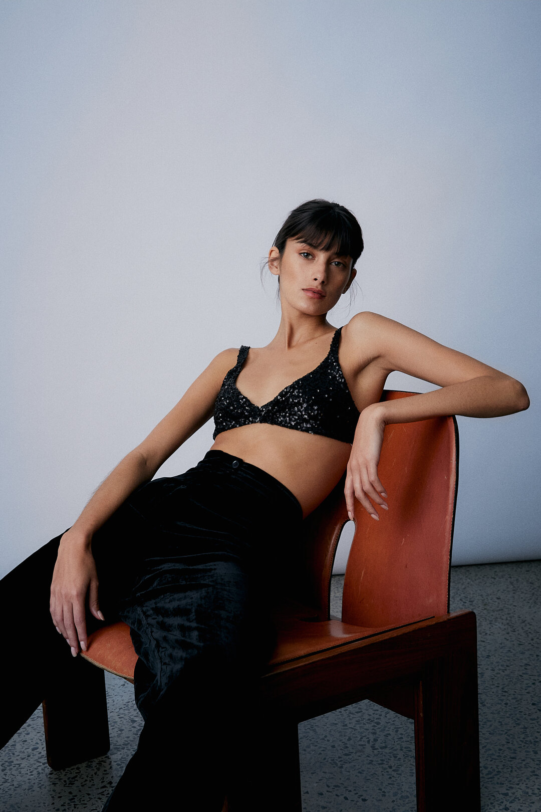 A brunette adult woman is seated on a wooden chair wearing a black sequined top and velvet pants in a brightly lit interior.