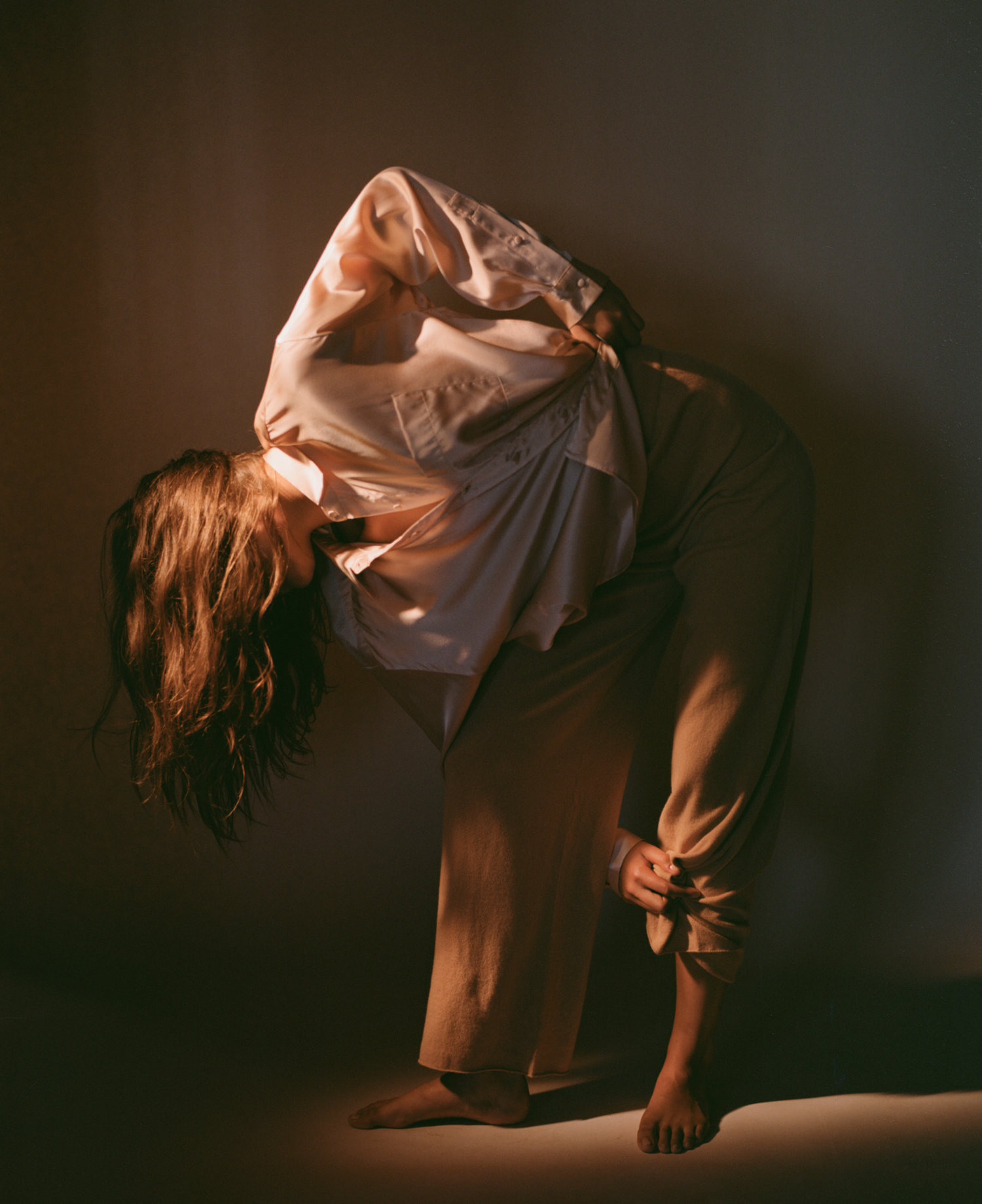 A person in a brightly lit, neutral-toned indoor setting, wearing loose clothing, with their head covered by a shirt and bare feet visible.