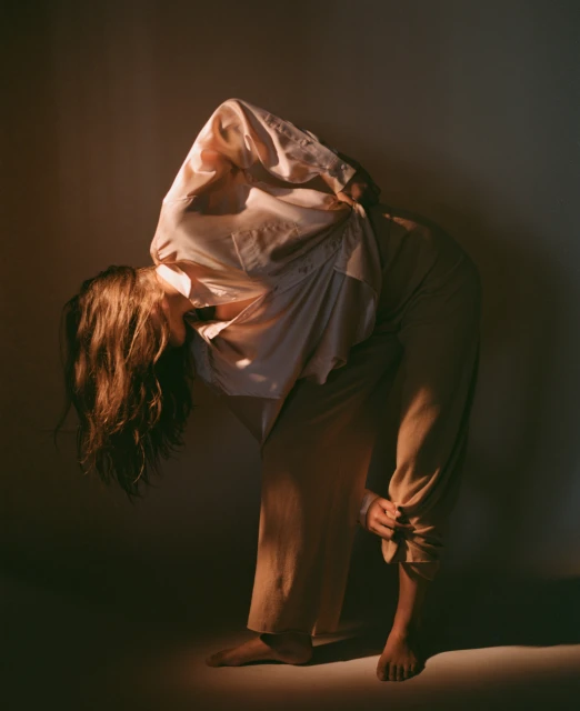 A person in a brightly lit, neutral-toned indoor setting, wearing loose clothing, with their head covered by a shirt and bare feet visible.