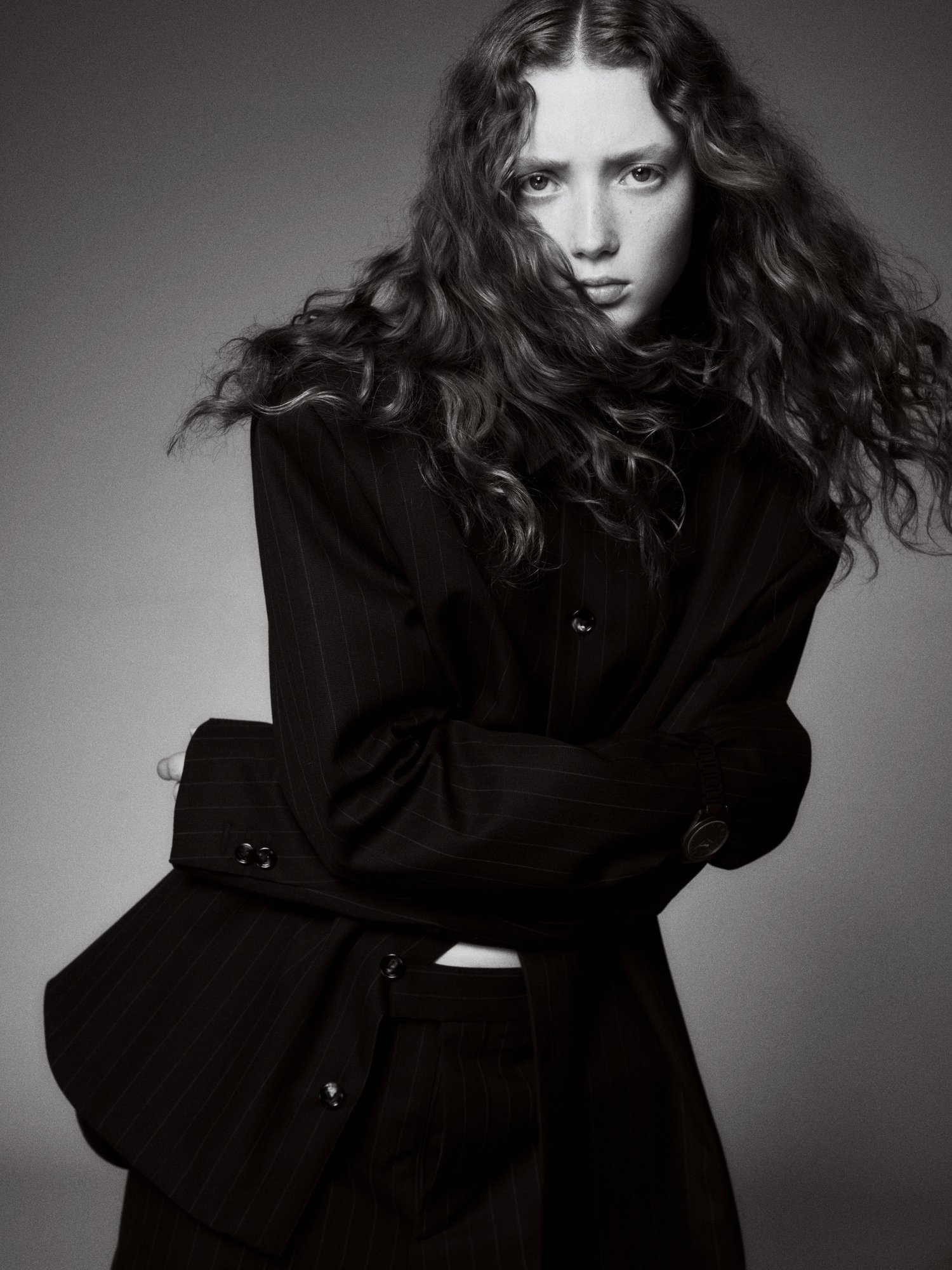 Monochrome portrait of a long-haired young woman in a dark pinstripe suit with arms crossed, set against a simple grey studio backdrop.