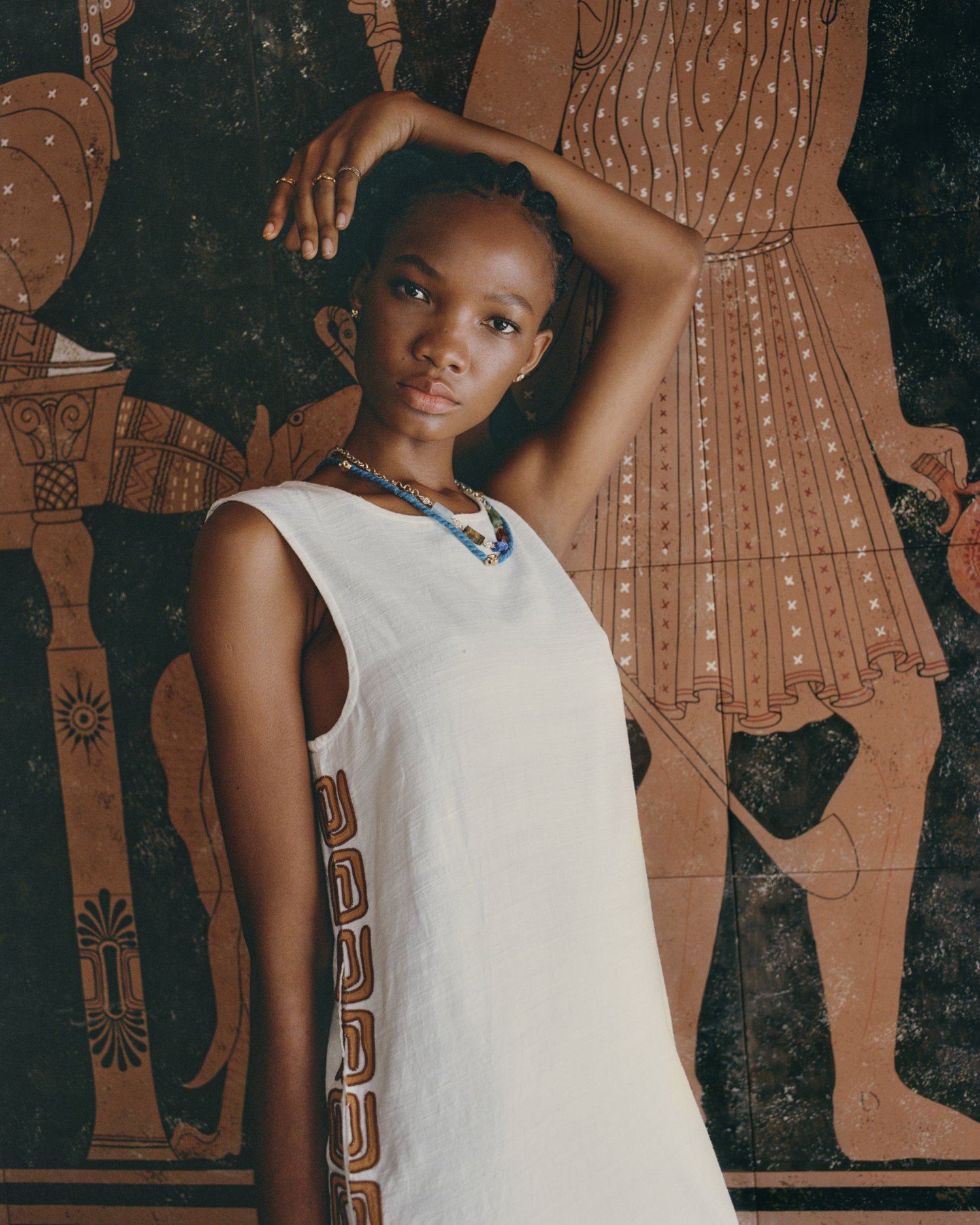 Portrait of Black woman in white dress posing in front of painted wall with classic figures and motifs in warm brown hues.