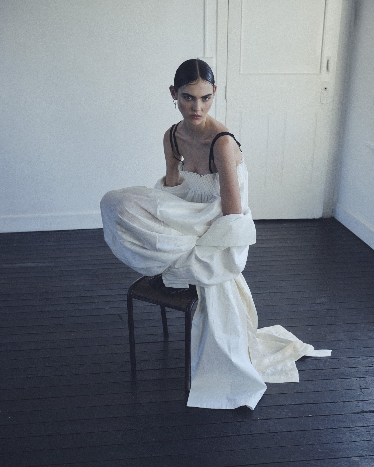 A woman in a white dress poses on a stool in a minimalist indoor setting, with a light and airy aesthetic.