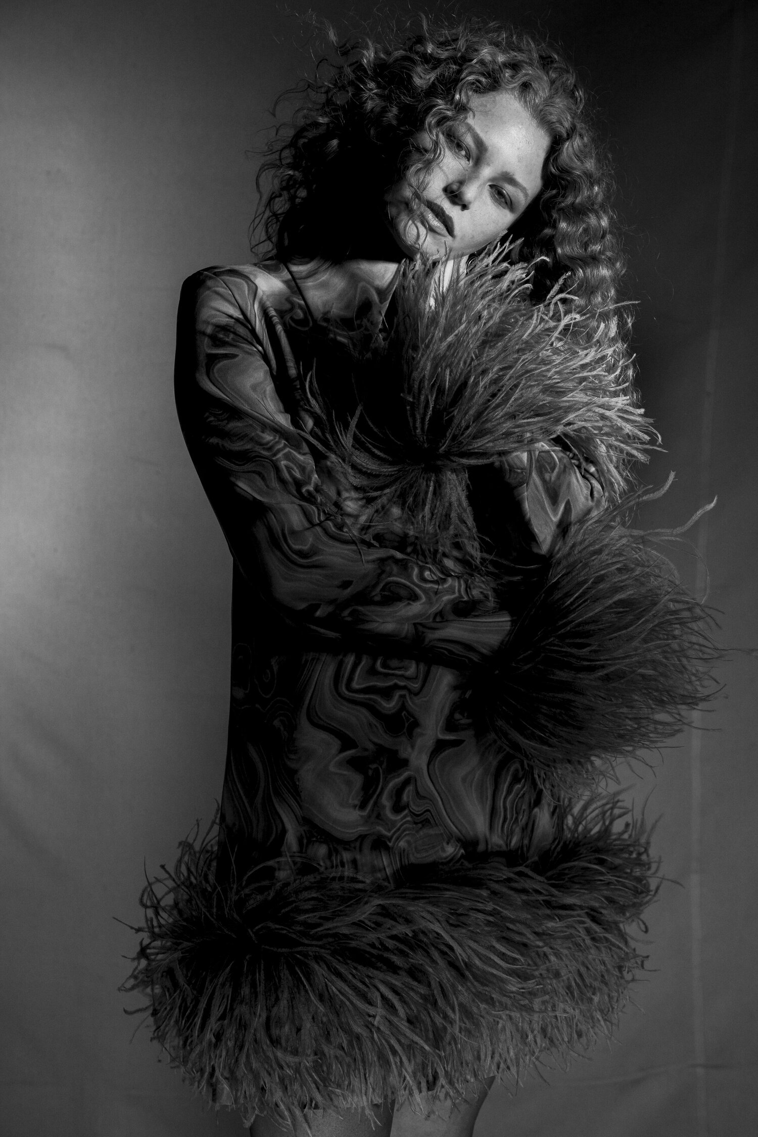 Black and white portrait of a young woman with curly hair wearing a short dress with feathered trim, posing in a photo studio.