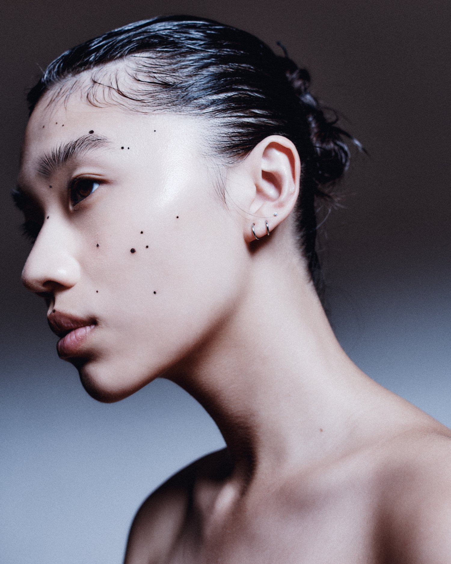 Close-up side portrait of a young Asian woman with slicked-back hair and artistic black dots on her face, set against a graduated gray backdrop.