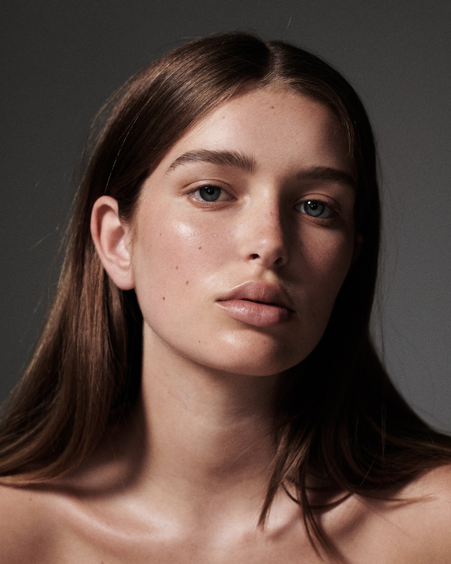 Portrait of a young woman with long brown hair, light skin, and blue eyes looking directly at the camera in a close-up studio shot.