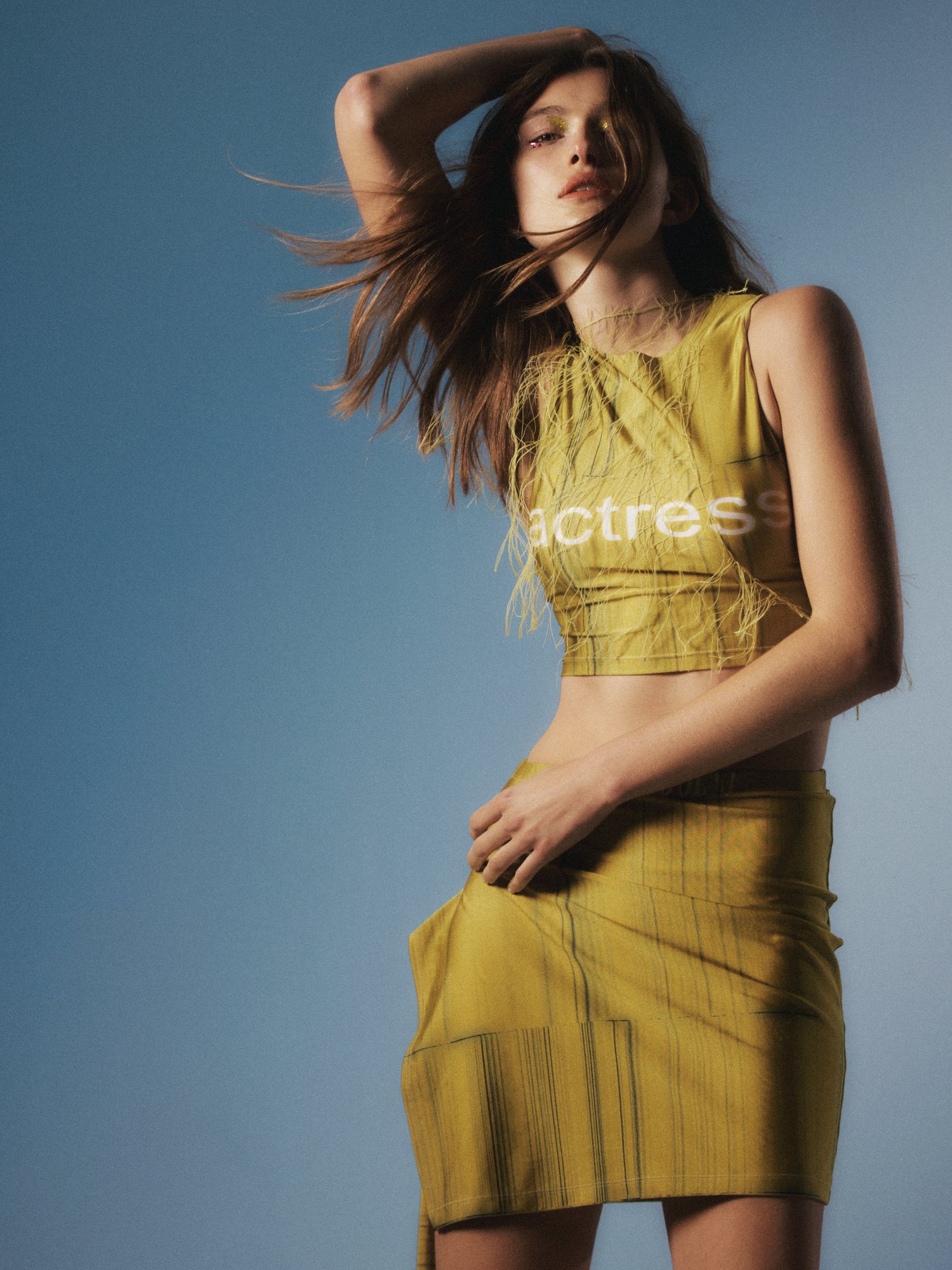 Young woman poses in a mustard-colored two-piece outfit, consisting of a crop top and skirt against a soft blue gradient background.
