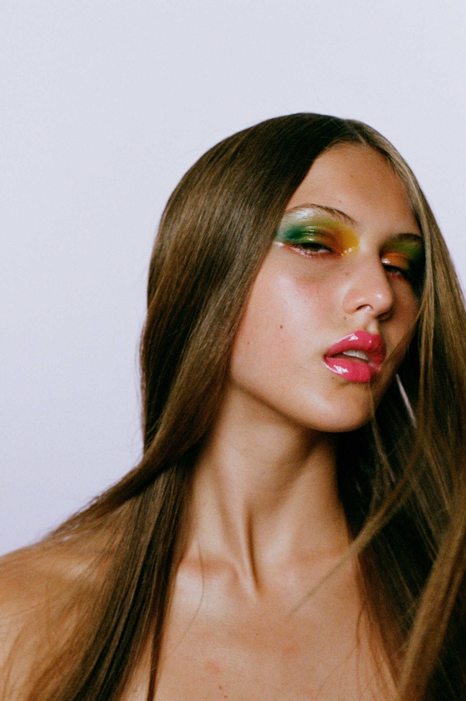 Close-up studio shot of a woman with long brunette hair, wearing multicolored eye makeup and glossy pink lipstick, against a white backdrop.
