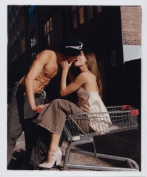 A shirtless man in a beanie and a woman in a cream slip dress kissing while she sits in a shopping cart in Sydney.