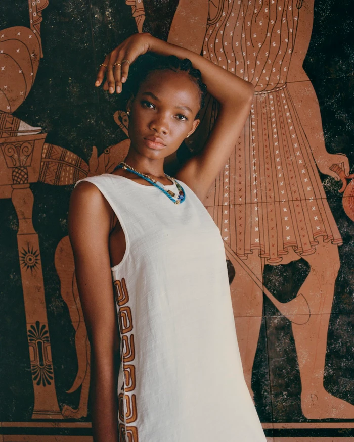 Black woman with braided hair wearing a white sleeveless dress with patterned side trim standing before a classical painted mural.