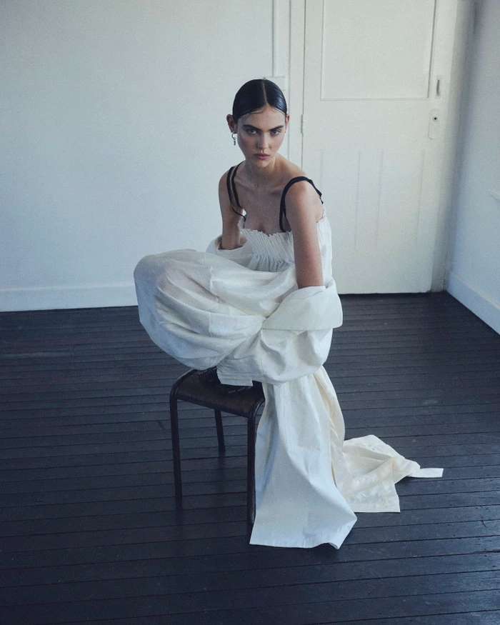 Model in a voluminous white dress crouched on a dark chair against a white wall.