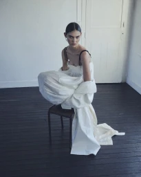 Model in a voluminous white dress crouched on a dark chair against a white wall.
