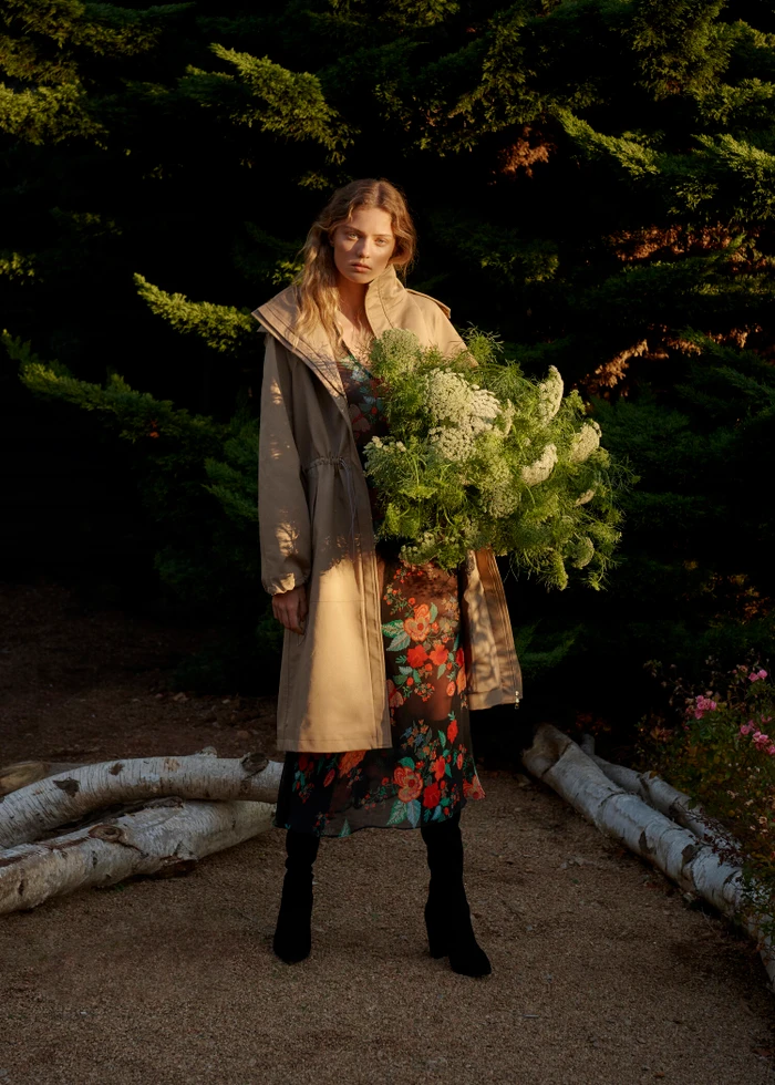 Model standing outdoors holding a large floral bouquet while wearing a tan hooded coat and floral dress.