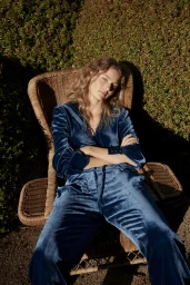 Woman in blue velvet loungewear seated in a wicker armchair outdoors with dappled sunlight against a dense green hedge.
