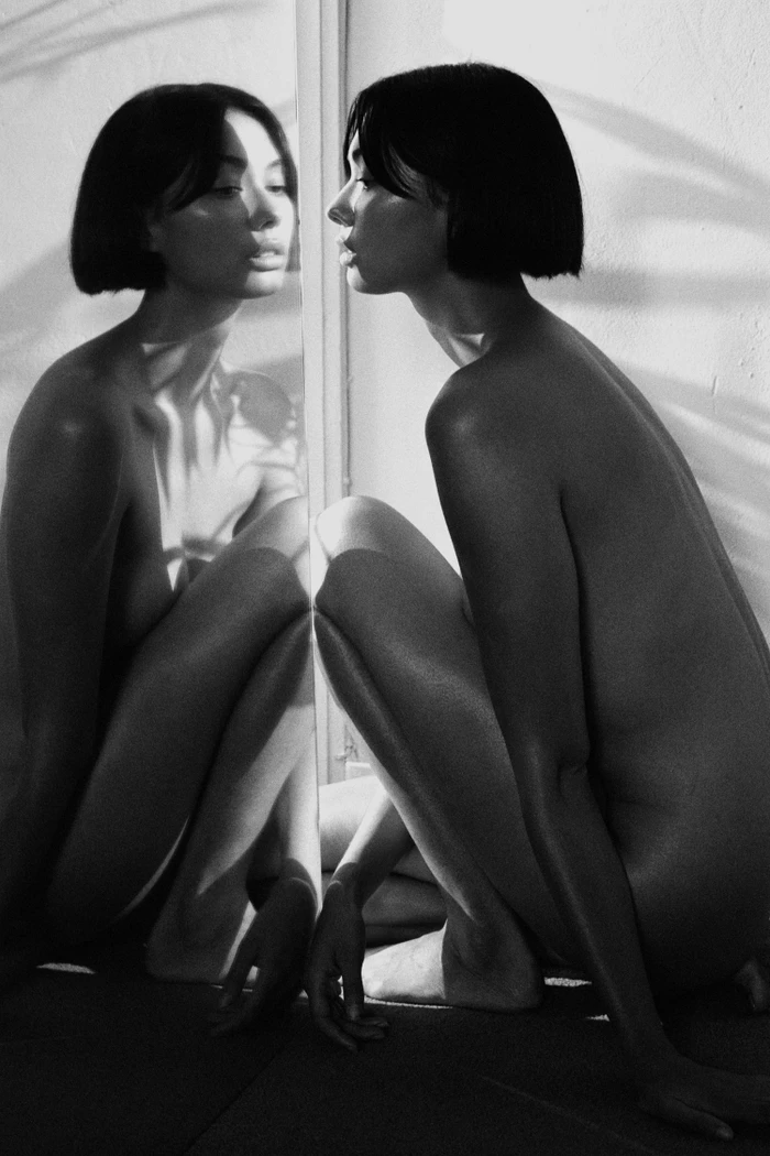 Black and white portrait of a woman with a sleek bob haircut crouching and looking into a mirror
