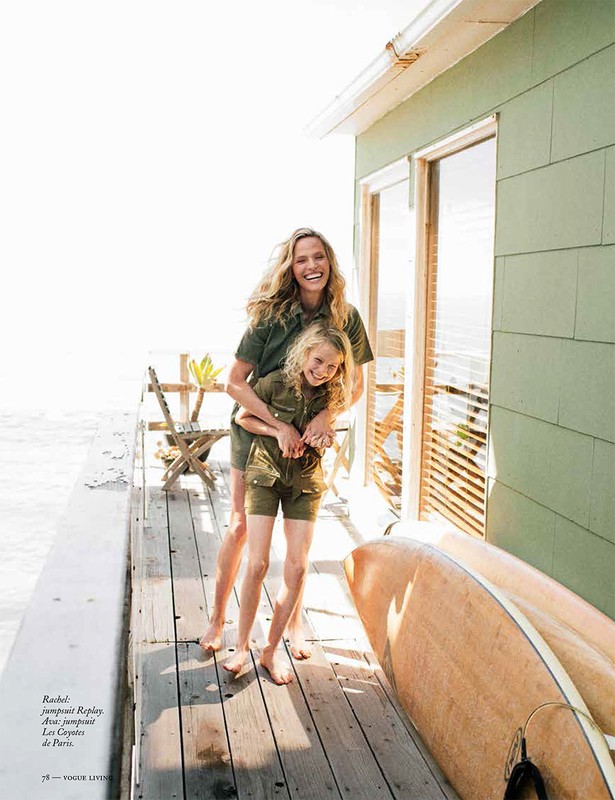 Mother and daughter in matching outfits embrace on a deck overlooking the ocean, with a surfboard and a coastal home nearby.