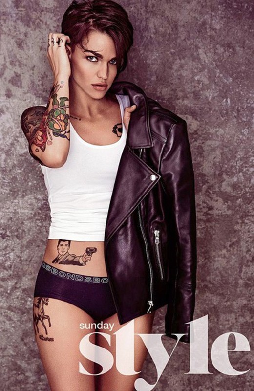 Ruby Rose wearing a white tank top and leather jacket with visible tattoos against a textured grey background.