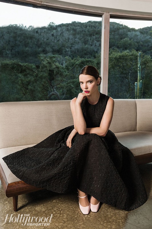 Anya Taylor-Joy sits on a sofa in a black textured gown against a natural backdrop. Soft daylight, medium shot, Hollywood Reporter logo visible.