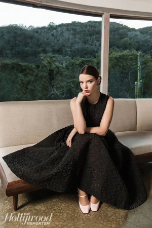 Anya Taylor-Joy sits on a sofa in a black textured gown against a natural backdrop. Soft daylight, medium shot, Hollywood Reporter logo visible.