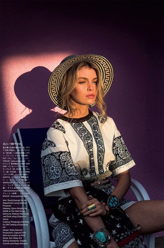 A blond woman in a straw hat sits in a white chair against a purple wall, wearing a patterned tunic and turquoise jewelry in outdoor light.