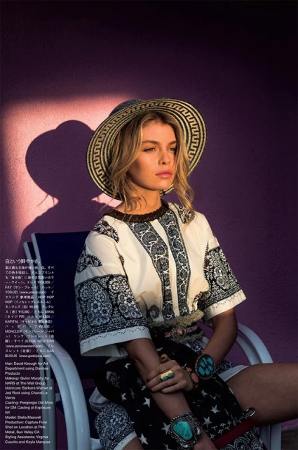 A blond woman in a straw hat sits in a white chair against a purple wall, wearing a patterned tunic and turquoise jewelry in outdoor light.