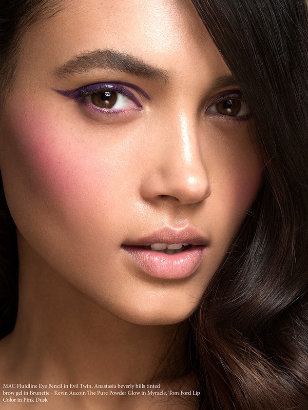 Close-up beauty shot of a young adult woman with purple eyeliner and pink blush, showcasing makeup in a studio environment.