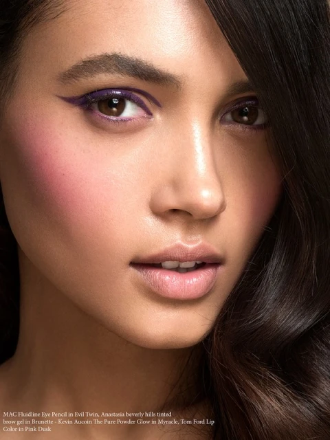 Close-up beauty shot of a young adult woman with purple eyeliner and pink blush, showcasing makeup in a studio environment.