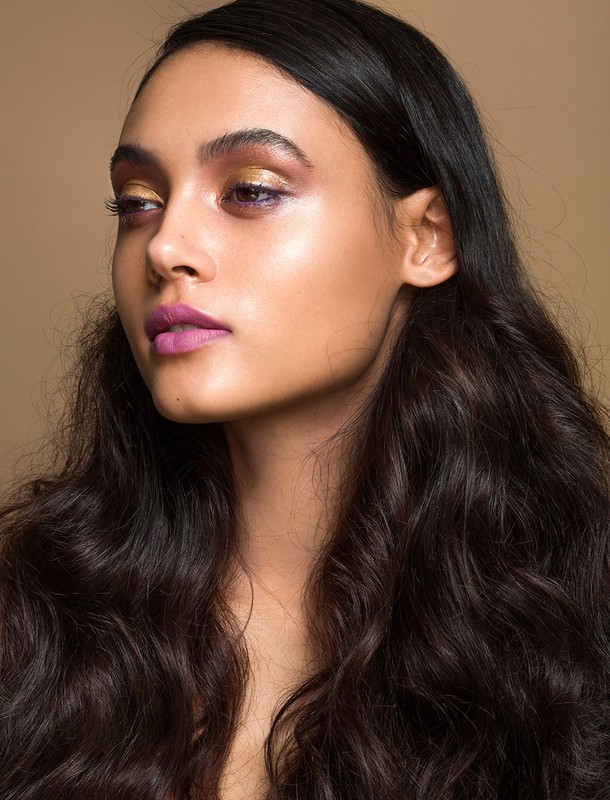 Close-up of a young adult female with dark wavy hair and professional makeup including gold eyeshadow and purple lipstick against a tan backdrop.