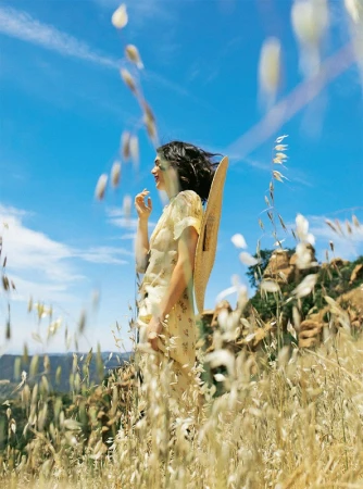 Woman in floral dress and straw hat standing in a tall grass field under a bright blue sky.