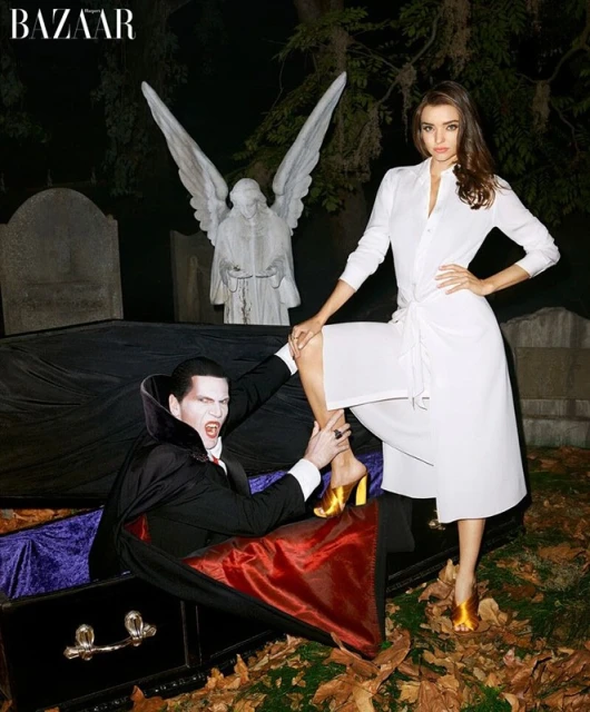 A woman in a white dress poses with a vampire rising from a coffin in a graveyard setting, featuring a gothic-inspired fashion editorial.