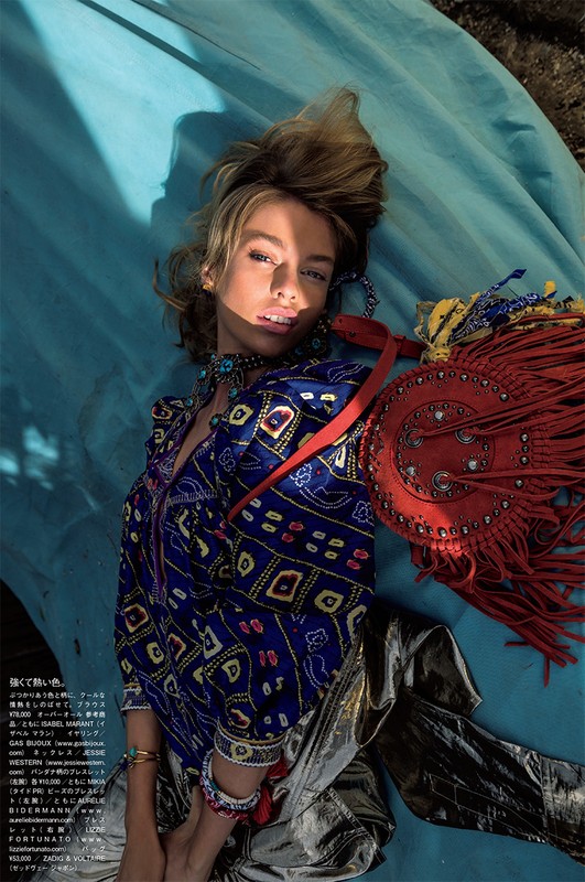 A woman poses in a blue patterned top and silver pants, lying on a blue backdrop with an orange fringed purse. Fashion shoot.