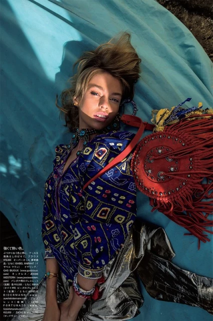 A woman poses in a blue patterned top and silver pants, lying on a blue backdrop with an orange fringed purse. Fashion shoot.