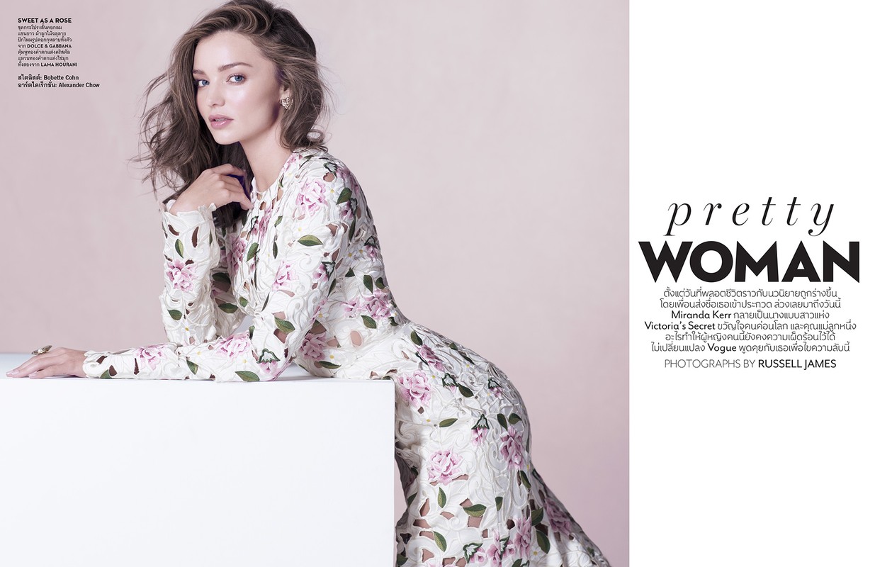 Miranda Kerr poses in a long-sleeved, floral-patterned dress against a soft pink backdrop. She is leaning on a white block in a fashion shot.