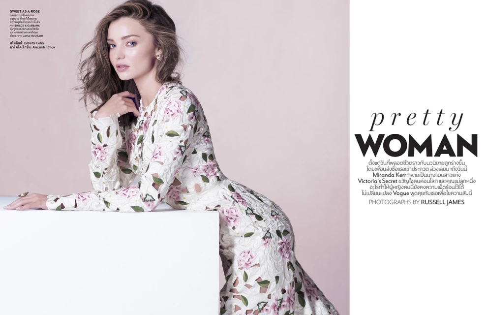 Miranda Kerr poses in a long-sleeved, floral-patterned dress against a soft pink backdrop. She is leaning on a white block in a fashion shot.
