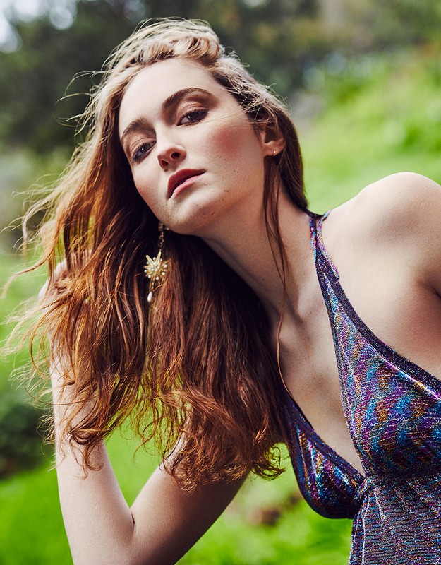 A woman with red hair poses outdoors wearing a shiny iridescent top and starburst earrings, set against a lush green background.