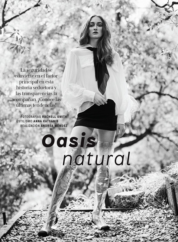 Black and white fashion shot of a model in a sheer white top, black skirt, and transparent boots posing in front of a natural background.