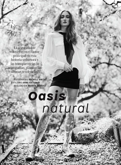 Black and white fashion shot of a model in a sheer white top, black skirt, and transparent boots posing in front of a natural background.