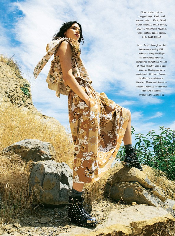 A woman in a floral top and skirt stands on rocks in a sunlit outdoor setting, wearing ankle boots; blue sky with clouds visible.