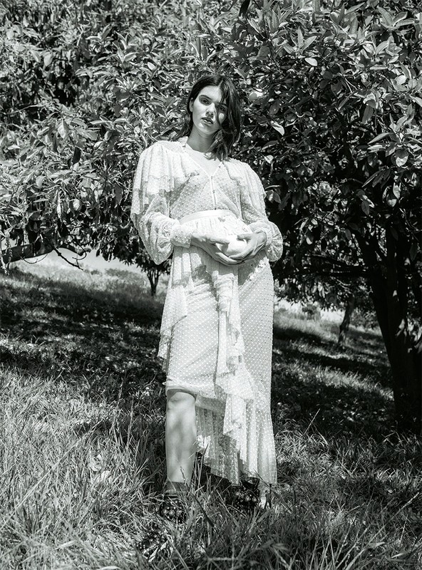 Monochrome portrait of a woman in a floral dress standing in a grassy field near a tree with dark leaves, creating a natural, serene setting.