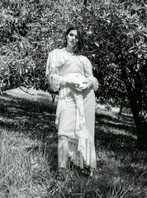 Monochrome portrait of a woman in a floral dress standing in a grassy field near a tree with dark leaves, creating a natural, serene setting.