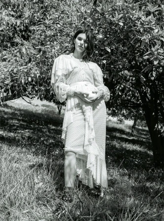 Monochrome portrait of a woman in a floral dress standing in a grassy field near a tree with dark leaves, creating a natural, serene setting.