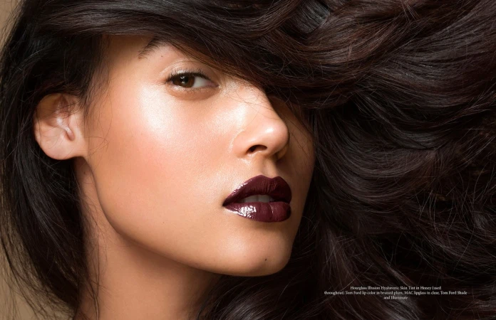 Close-up beauty portrait of a woman with glossy dark plum lipstick and voluminous dark brown hair falling across her forehead.