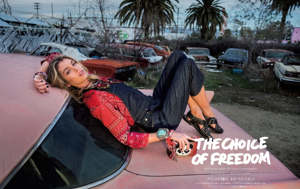 Fashion editorial photo of a blonde woman in denim overalls and a red patterned shirt, lying on a pink car in a junkyard with palm trees.