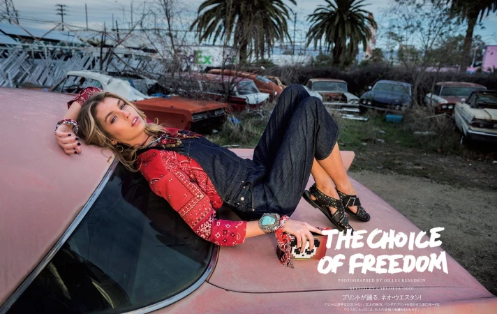 Fashion editorial photo of a blonde woman in denim overalls and a red patterned shirt, lying on a pink car in a junkyard with palm trees.