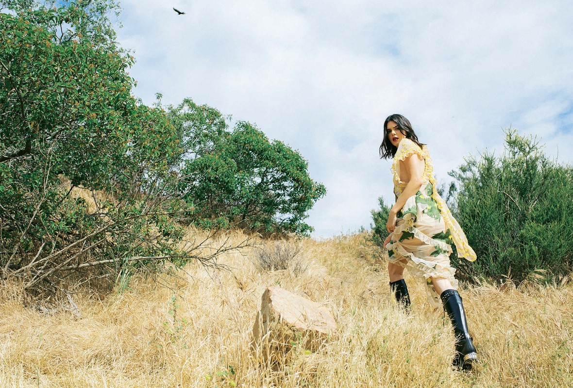 Woman in floral dress and black boots walks through golden field on hillside under cloudy sky, trees and bird visible.