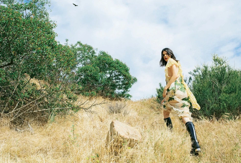 Woman in floral dress and black boots walks through golden field on hillside under cloudy sky, trees and bird visible.