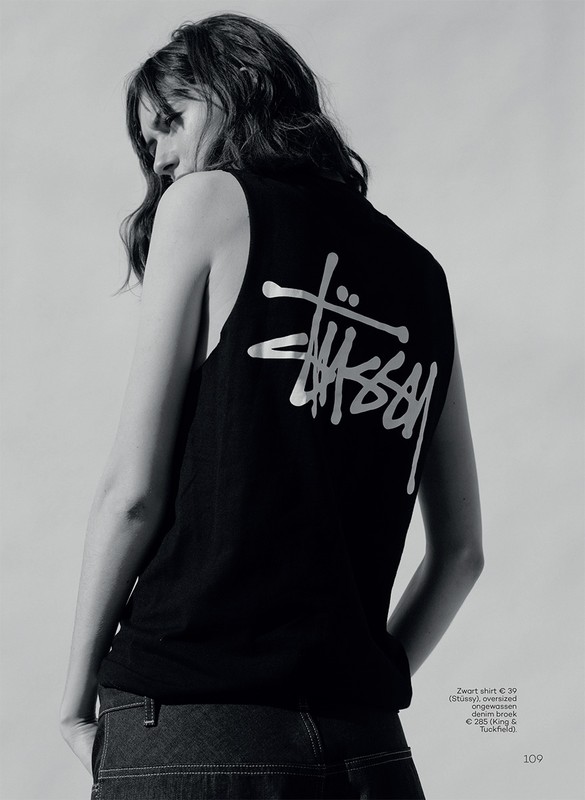An adult female wearing a black Stüssy tank top poses in a studio setting, captured in a medium shot with a monochromatic color scheme.